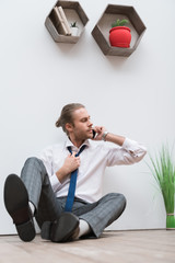 Businessman sitting on a wooden floor, talking by smartphone and loosening tie