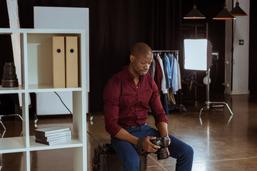 Fototapeta premium portrait of african american photographer looking at photo camera in hands while choosing photos in studio