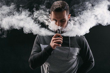 Young man vaping e-cigarette with smoke on black