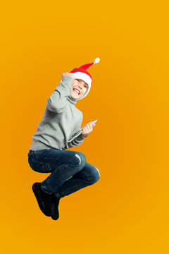 Excited Boy In Christmas Hat Jumping On Yellow Wall Background.