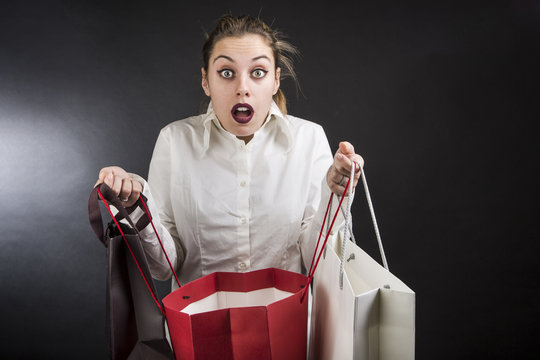 Giovane ragazza bionda con camicia bianca ci guarda con le buste per lo shopping tenute dietro aperte e fa un espressione di stupore  - sfondo nero