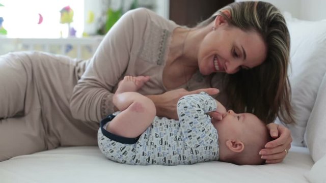 Young mother, lying in bed with her newborn baby boy, playing and interacting together. Family happiness concept. Back light fron the window behind