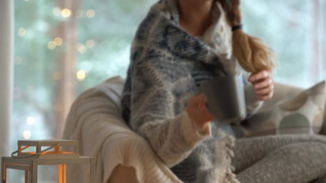 Cozy winter lifestyle. Young happy woman drinking cup of coffee wearing knitted sweater sitting home by the big window with winter snow tree background