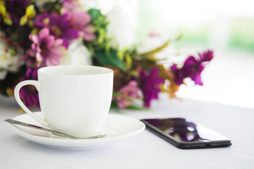 White cup of black coffee, smartphone and flower placed on white table background at coffee shop.