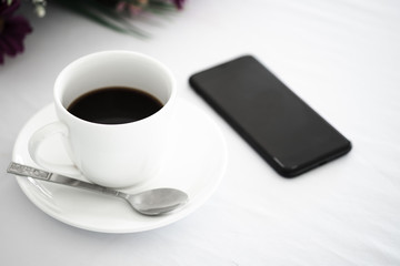 White cup of black coffee, smartphone and flower placed on white table background at coffee shop.