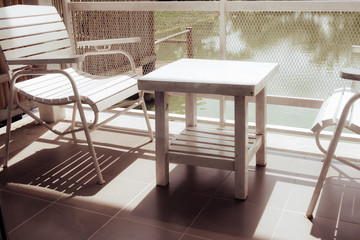 White wooden table and chairs are located on terrace of house and lake view with white steel fence and sun shining in morning. Balcony seating for relaxation in vintage tone.