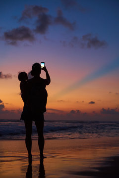 silhouette of young mother with child making photo of sunset by smartphone