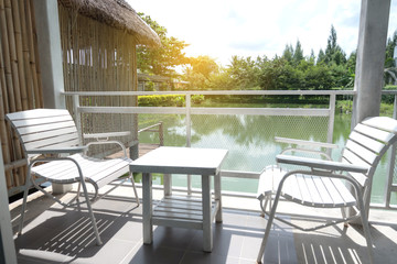 White wooden table and chairs are located on terrace of house and lake view with white steel fence and sun shining in morning.