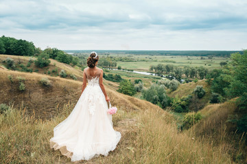 A young girl, the bride in a long wedding dress, is turned her back and stares into the distance to the river and a beautiful landscape.