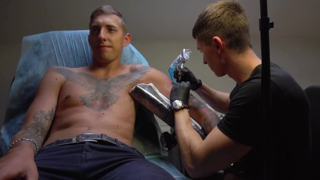 Guy smiles sitting in the chair while the master makes him a tattoo on his arm