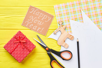 Fototapeta premium New Year handmade background, top view. Cut out wooden decoration deer, scissors, pencil, gift box, message on yellow wooden background. Happy New Year.