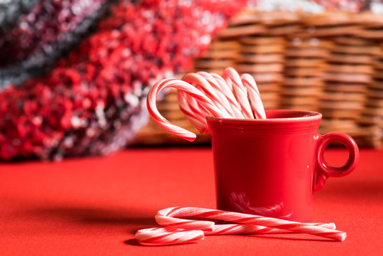 A Red Mug Holding Christmas Candy Canes On A Red Tabletop With Two Candy Canes In Front And A Basket Holding A Knitted Afghan In The Background.