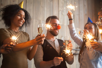 African and caucasian friends holding sparklers celebrating New year party, diverse people in funny hats hanging out together with champagne glasses on holiday eve, multiracial guys enjoy celebration