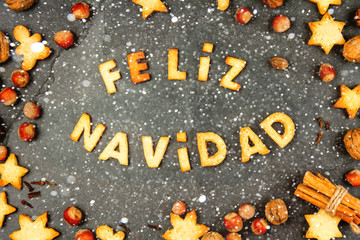 FELIZ NAVIDAD COOKIES. Words Merry Christmas en Spanish with baked cookies, Christmas decoration and nuts on black slate background. Christmas card for hispanic countries top view. snow effect