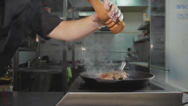 Meat frying on griddle pan. Crop of chef pouring spices in cooking steak, at restaurant commercial kitchen, slide slow motion