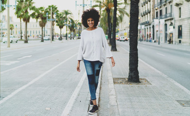 Full length portrait of smiling student female in casual clothing while standing on urban setting copy space area for advertising, young woman with curly hair enjoying city walking.