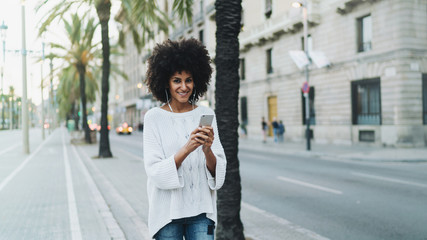 Young hipster girl communicationg with friend while walking the city with mobile phone. Smiling hipster girl chatting online while relaxing outdoors with a music downloaded and played on a smartphone.