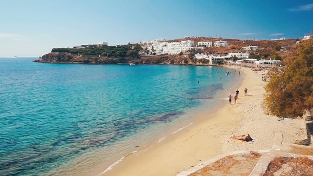 Colorful sandy beach with azure water, tourists and local white buildings. Agios Stefanos Beach of Mykonos island, Greece.