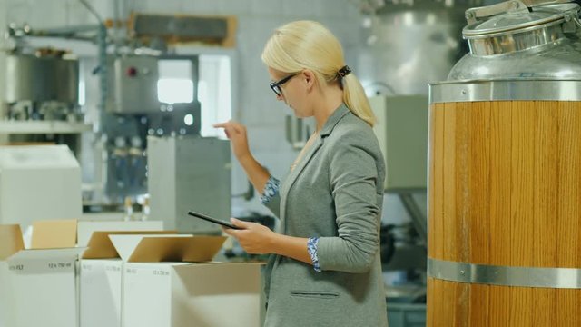 A woman with a tablet makes an inventory in the finished goods warehouse. Nearby there are boxes of wine and a large cistern