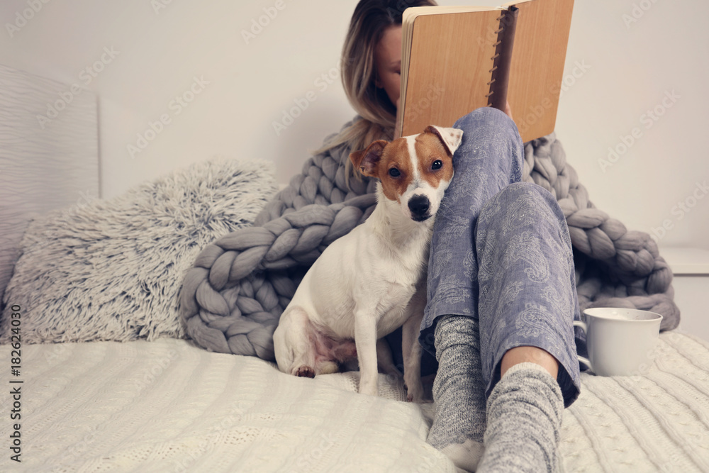 Wall mural Woman in cozy home clothes relaxing at home with dog Jack Russel Terrier, drinking cacao, reaing a book. Comfy lifestyle.