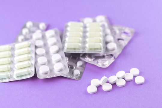 Full and empty packs of white capsules and pills packed in blisters with copy space on purple background. Focus on foreground, soft bokeh