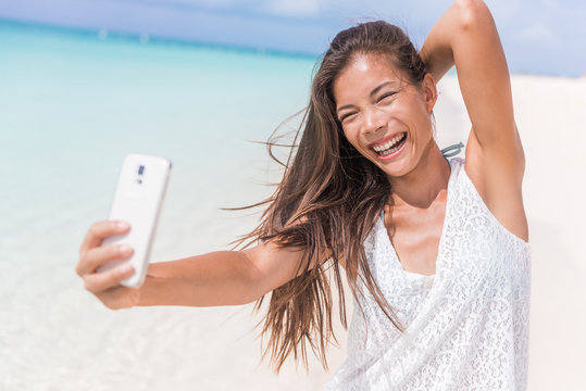 Selfie summer beach vacation fun girl laughing. Happy Asian woman taking self-portrait pictures with mobile phone on tropical holidays in Caribbean posing for camera.