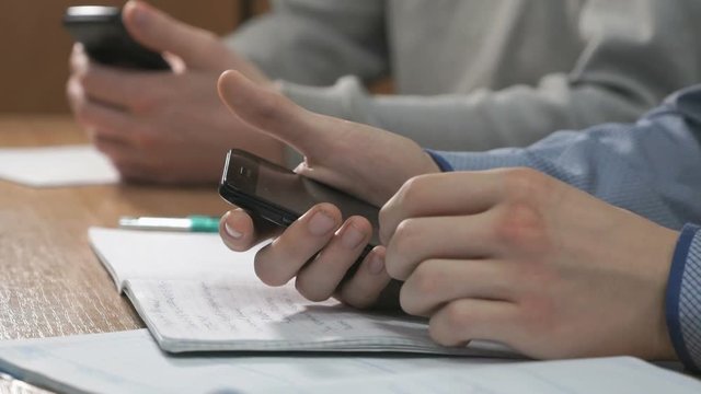 Close-up of hands of two unknown students dressed in shirt sits at desk at lesson and using black smartphones pressing them fingers on the screen and look at the important information.