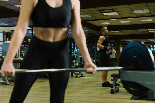 Close up - body sport woman. Sporty woman exercising with barbell in gym. Strong man with dumbbells in the background