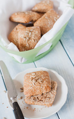 Homemade square breakfast bread rolls made from whole grain flour, sunflour seeds, sesame seeds and various other seeds and grains in bread basket standing at light blue wooden table with vintage b