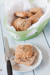 Homemade square breakfast bread rolls made from whole grain flour, sunflour seeds, sesame seeds and various other seeds and grains in bread basket standing at light blue wooden table with vintage b