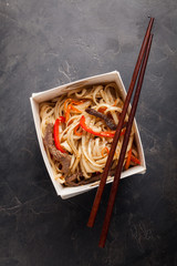 Close-up of noodles in a box with vegetables and beef in teriyaki sauce. Top view of Asian food, and wooden sticks. The concept of fast food