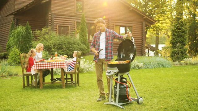 Handsome father grilling vegetables on the barbecue while his family sitting at the table behind him. Pictutesque green garden with a modern wooden home. Outside