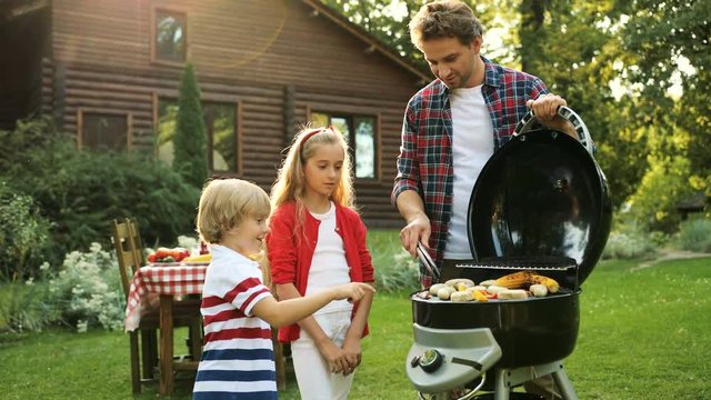 Handsome father grilling on the barbecue and his kids watching him. Beautiful green garden and wooden modern house on the backgrond. Outside