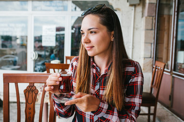 Beautiful young girl with sunglasses on head sitting at table in an outdoor cafe and drinking tea from glass beaker
