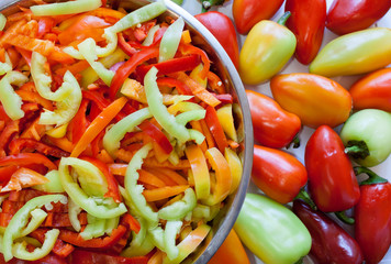 Sliced lengthwise stripes of multi-colored peppers are lying in round metal bowl and whole uncut peppers on table.