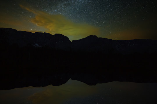 Stars in the night sky above the mountains and the lake. The Milky Way is reflected in the water surface.