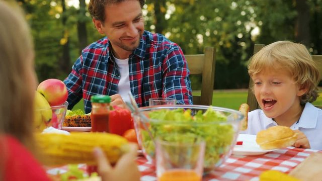 Close up of handsome father and son sitting at the table and boy biting a sausage in the green garden. Outdoor
