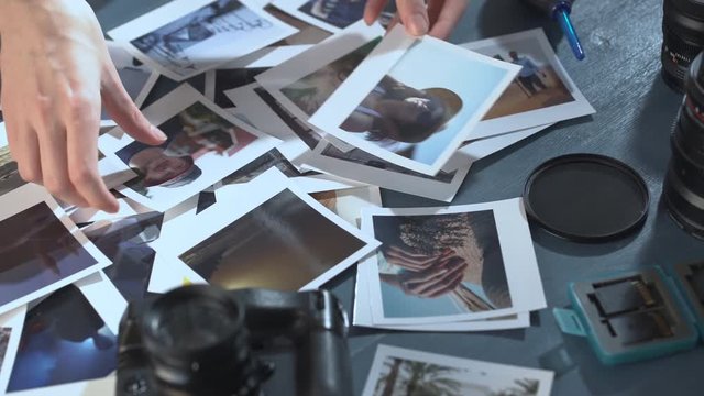A woman looks at printed photos from travel with sea and sun, spread on a wooden table with photo camera and lens.
