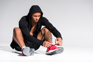 young african american sportsman in hoodie sitting and tying shoelaces on grey