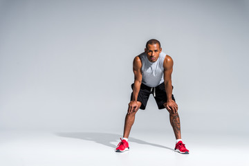 full length view of athletic young african american sportsman standing with hands on knees and looking at camera on grey