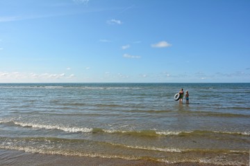 Zwei Kinder von hinten, stehen mit einem Schwimmreifen im Meer, Wellen im Vordergrund