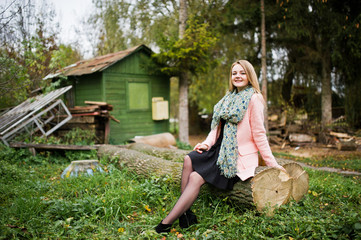 Young blonde girl at pink coat posed on autumn park, sitting on cut tree against house of the forester.