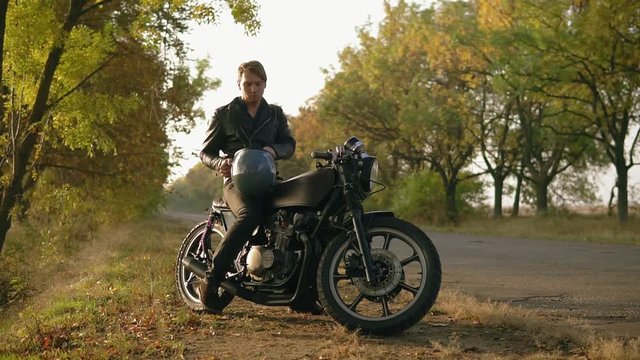 A young man in black leather jacket comes up to his motorcycle, sits there and puts a black helmet and sunglasses before starting his journey in the forest. Slow motion shot