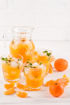 Mandarin cocktail with ice and mint in beautiful glasses and jug, fresh ripe citrus on white wooden background. Sweet orange juice. Close up photography. Selective focus