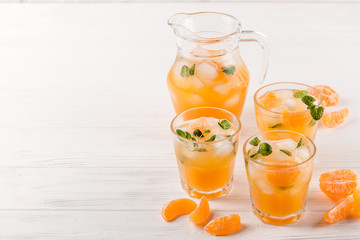 Mandarin cocktail with ice and mint in beautiful glasses and jug, fresh ripe citrus on white wooden background. Sweet orange juice. Close up photography. Selective focus