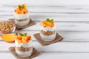 Obraz na płótnie Canvas Persimmon creamy trifle in beautiful glasses, fresh ripe fruit slices on white wooden background. Healthy vegetarian food. Delicious dessert. Close up photography. Selective focus.
