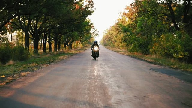 Front view of stylish man in black helmet and leather jacket riding motorcycle on an asphalt road on a sunny day in autumn. Beautiful trees with yellow and brown leaves around the road