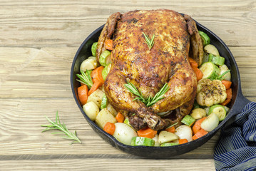 Roasted whole chicken with fresh vegetables and rosemary in black metal pan placed on rustic wooden background. Top view with copy space. Slow food concept