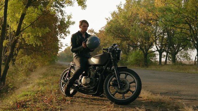 Attractive man in black leather jacket comes up to his motorcycle, sits there and puts a black helmet and sunglasses before starting his journey in the forest