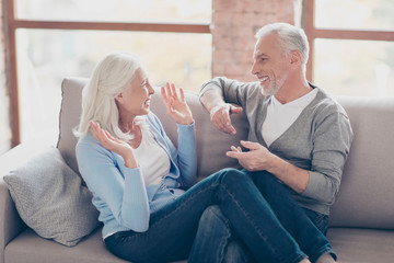 Happy senior couple having fun, looking to each other, laughing, sitting in living room, woman's legs lying on man's knees, couple expressing their emotion with hands